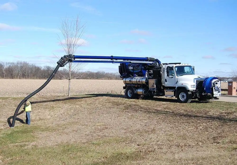 A white industrial vacuum truck with a long blue articulated arm and black suction hose is parked in a field, operated by a worker in a yellow safety vest.