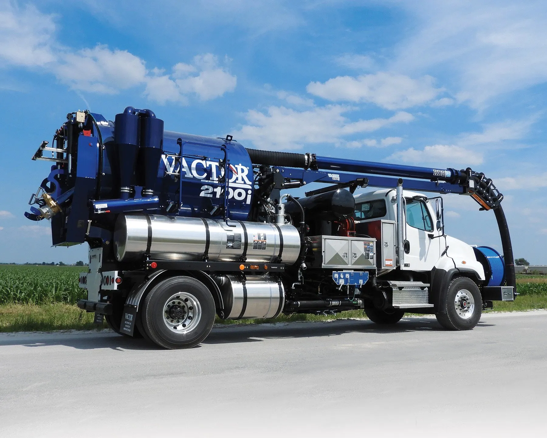 A blue and white Vactor 2100i vacuum truck parked on a road with a green field in the background under a partly cloudy sky.