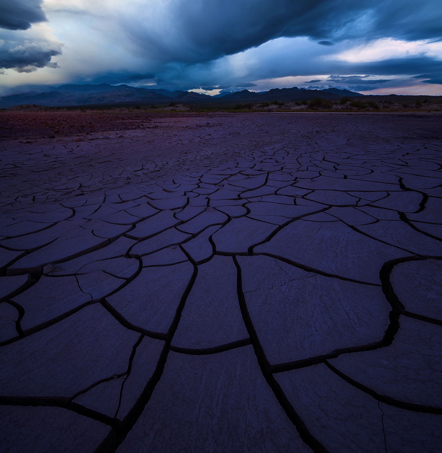 A vast, cracked desert floor stretches toward distant mountains under a dramatic, stormy sky.